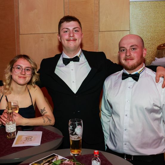Drei junge Erwachsene, zwei Männer und eine Frau, stehen gemeinsam an einem Tisch. Die Frau trägt ein schwarzes Kleid, und die Männer tragen Smokings. Sie lächeln und scheinen für ein Foto zu posieren.