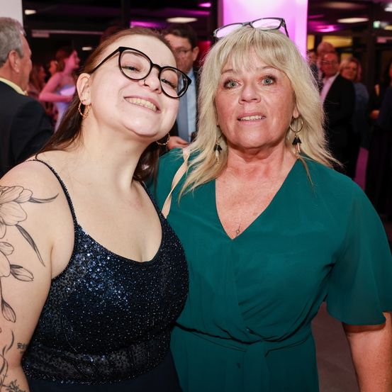 Zwei Frauen stehen eng beieinander bei einer formellen Veranstaltung, lächeln und posieren für ein Foto. Die Frau auf der linken Seite hat ein Blumentattoo auf der Schulter. Sie sind von anderen Menschen umgeben.