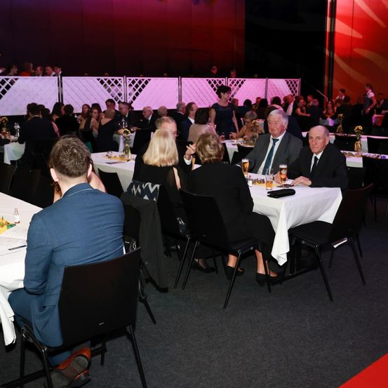 Eine Gruppe von Menschen sitzt an Tischen in einem Bankettsaal. Einige unterhalten sich, während andere ihre Mahlzeiten zu genießen scheinen. Der Raum ist mit einem rot-weißen Hintergrund dekoriert.