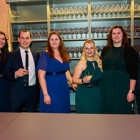 Fünf Menschen stehen lächelnd vor einem Regal voller Weingläser, zwei mit Gläsern in der Hand. Ein Mann im Anzug, eine Frau in einem blauen Kleid, eine weitere Frau in einem grünen Kleid und zwei weitere Frauen.