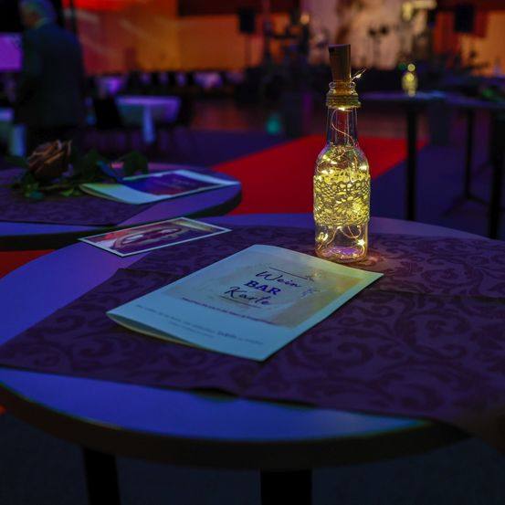 Eine Flasche mit Lichterketten steht auf einem mit einem lila Tuch bedeckten Tisch, mit einer Menükarte mit dem Titel 'Wein Bar Karte'. Im Hintergrund ein schwach beleuchteter Raum mit einem roten Teppich und einer Person, die steht.