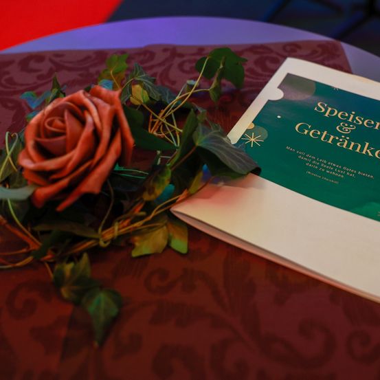 Eine rote Rose und grüne Blätter liegen auf einem Buch mit dem Titel 'Speiser & Getranke' auf einer gemusterten Tischdecke. Der Hintergrund ist unscharf und farbenfroh.
