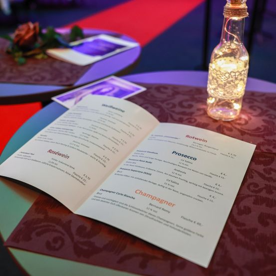 Ein Weinmenü liegt auf einem Tisch, mit einer Lichtflasche im Hintergrund. Das Menü enthält Optionen wie Roséwein, Prosecco und Champagner. Preise sind für jede Option aufgeführt.