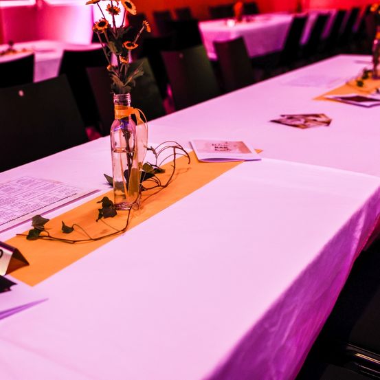 Ein langer Tisch mit weißer Tischdecke, einer Vase mit gelben Blumen und einem gelben Tischläufer. Papiere und Karten sind auf dem Tisch verstreut. Stühle sind um den Tisch herum angeordnet.