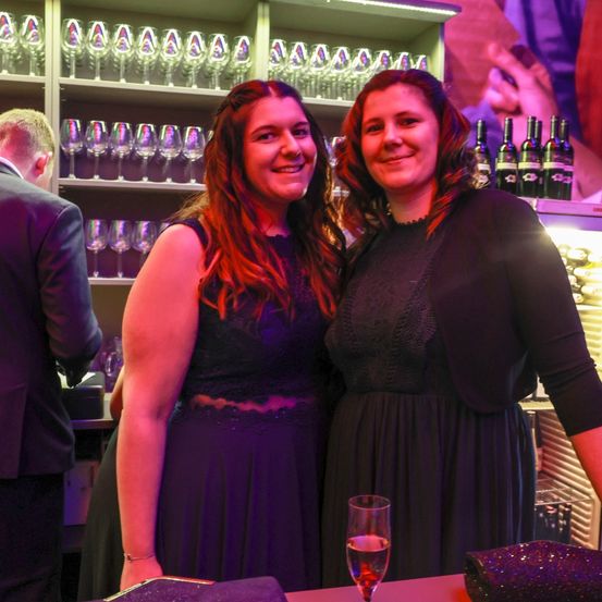 Zwei Frauen stehen lächelnd an einer Bar, eine hält ein Glas Wein. Hinter ihnen ist ein Mann in einem Anzug zu sehen. Im Hintergrund befinden sich Regale mit Weinläsern und Flaschen.