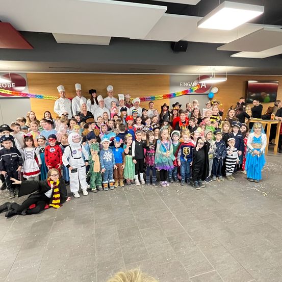 Eine große Gruppe von Kindern in Kostümen posiert für ein Foto, mit Köchen im Hintergrund. Ein bunter Banner ist sichtbar, und es gibt Luftballons und einen Tisch mit Gegenständen.