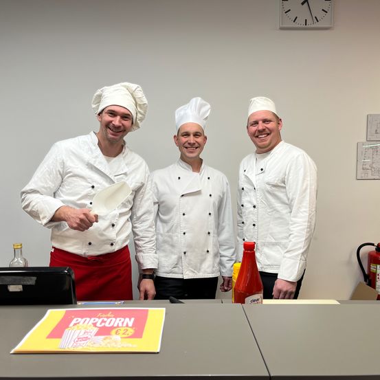 Drei Köche in weißen Uniformen und Hüten lächeln für ein Foto. Ein Menükarten und Gewürze sind auf dem Tisch. Eine Uhr und ein Feuerlöscher sind an der Wand montiert.