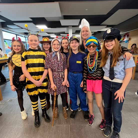 Eine Gruppe von Kindern und Erwachsenen in Kostümen posiert für ein Foto in einem Raum. Die Kinder tragen verschiedene thematische Kostüme, darunter eine Biene, eine Hexe und eine Polizistin. Eine erwachsene Frau in einer Kochmütze steht hinter ihnen.