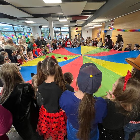 Eine große Versammlung von Kindern und Erwachsenen in einer Halle, die einen bunten Fallschirm beobachten. Viele sind in Kostümen gekleidet und einige tragen Hüte. Die Kinder stehen um den Fallschirm herum.
