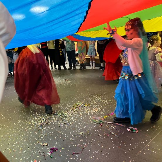 Eine Gruppe von Kindern in Kostümen tanzt und wirft Konfetti in einem Saal. Ein Kind in einem roten Kleid hält einen großen Regenschirm.