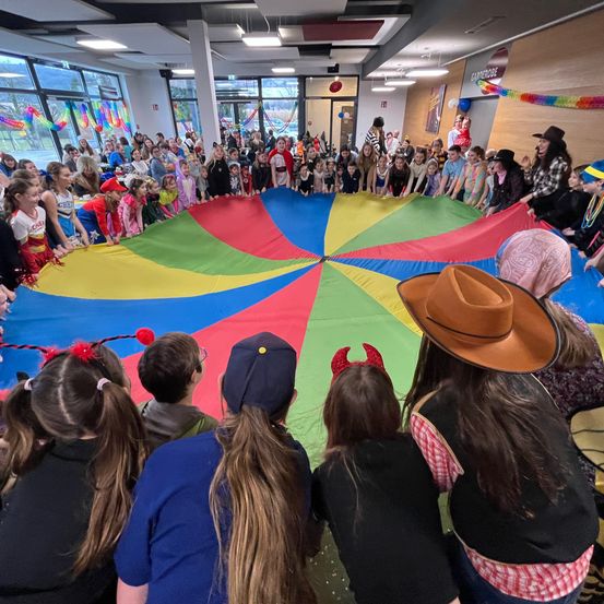 Eine Gruppe von Kindern in verschiedenen Kostümen versammelt sich um einen großen, bunten Fallschirm in einem Raum mit großen Fenstern. Der Fallschirm wird von mehreren Erwachsenen gehalten, und die Kinder schauen erwartungsvoll zu.
