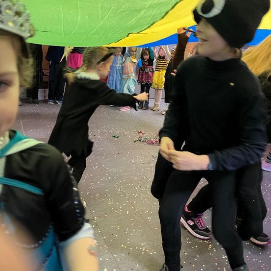Kinder in Kostümen tanzen unter einem großen grünen Blatt in einer festlichen Umgebung mit Konfetti auf dem Boden.