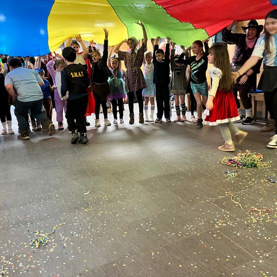 Eine Gruppe von Kindern und Erwachsenen hält einen farbenfrohen Fallschirm in einem Raum mit Konfetti auf dem Boden hoch. Einige Kinder tragen Kostüme.
