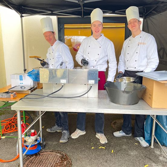 Drei Köche stehen unter einem Zelt und lächeln. Sie tragen weiße Uniformen und Kochmützen. Einer hält einen Schöpflöffel, ein anderer einen Pfannenwender und der dritte hat eine Schüssel. Ein Tisch davor hat verschiedene Kochgeräte.