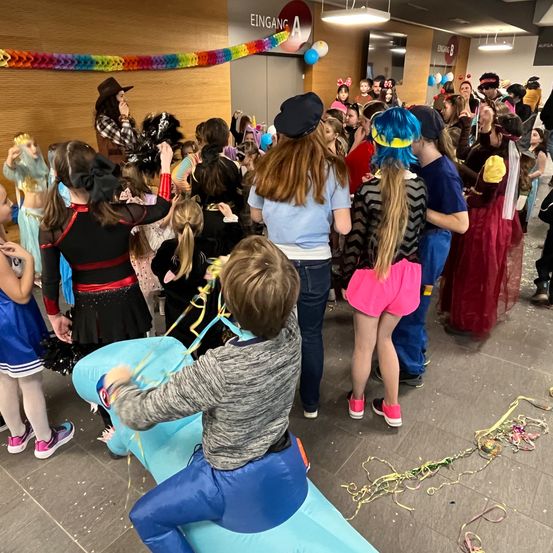 Eine Gruppe von Kindern in verschiedenen Kostümen, einige mit Hüten, sind drinnen versammelt. Sie stehen herum, manche sitzen auf einem blauen Luftkissen und scheinen an einer Aktivität beteiligt zu sein. Eine Frau in einem Cowboyhut steht vor ihnen.