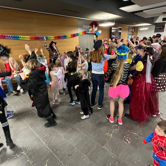 Eine große Gruppe von Kindern in Kostümen tanzt in einem Gebäude mit Erwachsenen. Ein bunter Banner ist über der Spitze des Raumes.