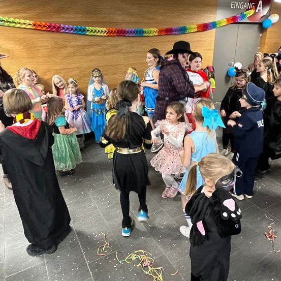 Eine Gruppe von Kindern in Kostümen versammelt sich in einem Raum, einige mit Masken. Ein Mann in einem Cowboyhut steht in der Mitte. Konfetti liegt auf dem Boden verstreut. Ein Schild über der Tür steht 'EINGAN A'.