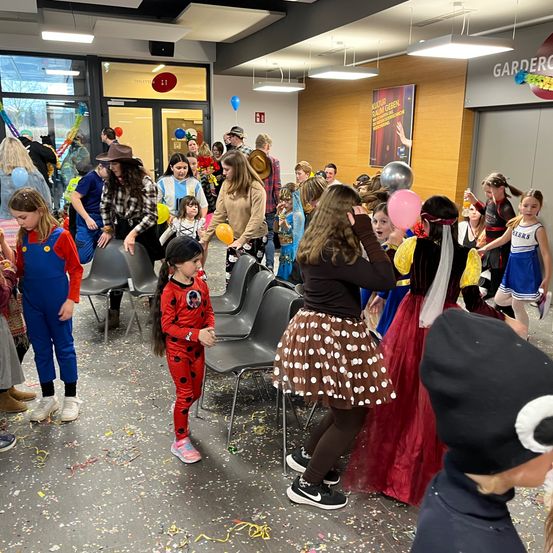 Eine Gruppe von Kindern und Erwachsenen versammelt sich in einem Raum für eine Party, trägt Kostüme, hält Luftballons und steht vor Stühlen. Der Boden ist mit Konfetti bedeckt, und zwei Personen stehen vor einer Tür.