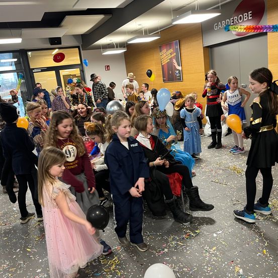 Eine Menschenmenge aus Kindern und Erwachsenen, einige in Kostümen, versammelt sich drinnen. Konfetti und Luftballons sind auf dem Boden verstreut. Einige sitzen, andere stehen. Eine Person in einer Polizeiuniform steht vorne.