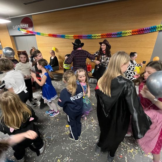 Eine Gruppe von Kindern in Kostümen, darunter ein Polizist, tanzt in einem Raum, der mit Luftballons und Wimpelketten dekoriert ist. Zwei Erwachsene, einer in einem Cowboyhut, befinden sich in der Mitte.