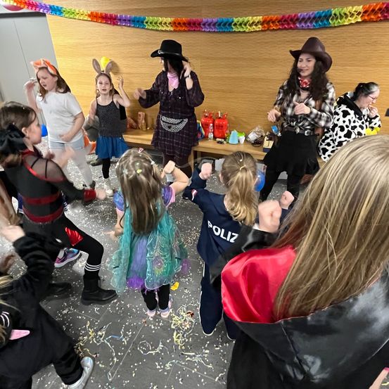 Eine Gruppe von Kindern in verschiedenen Kostümen tanzt in einem Raum, angeführt von zwei Frauen in Cowgirl-Kleidung. Dahinter befindet sich eine Holzwand mit einer bunten Papiergirlande und einem Tisch mit verschiedenen Gegenständen darauf.