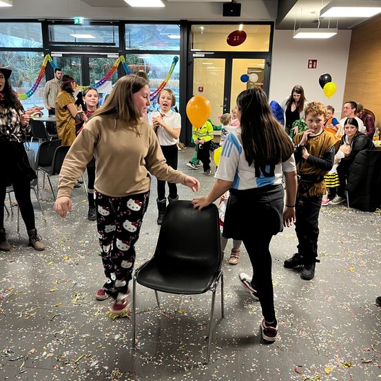 Eine Gruppe von Menschen ist in einem Raum mit Luftballons, Konfetti und Dekorationen versammelt. Zwei Frauen interagieren spielerisch mit einem schwarzen Stuhl, während andere zuschauen.