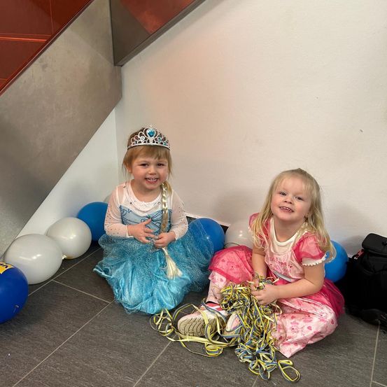 Zwei kleine Mädchen sitzen auf dem Boden in Prinzessinnenkleidern. Ein Mädchen ist als Elsa aus Die Eiskönigin verkleidet und das andere als rosa Prinzessin. Sie sind von Luftballons umgeben.