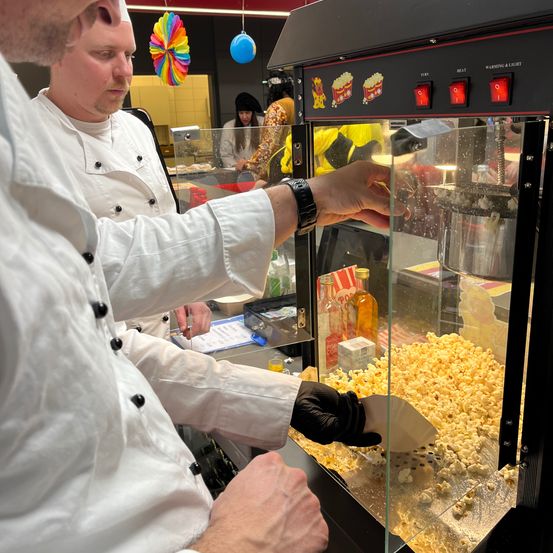 Ein Koch in weißer Uniform schaufelt Popcorn aus einer Maschine. Ein weiterer Koch steht hinter ihm. Die Maschine hat eine Glasfront und auf dem Tresen befinden sich Flaschen und andere Gegenstände.