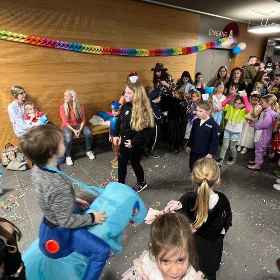 Eine Gruppe von Kindern in verschiedenen Kostümen versammelt sich in einem Flur. Einige tragen Hüte und Schuhe. Eine Erwachsene hält ein Baby, während andere zusehen. Ein blauer pferdeähnlicher Ballon und Konfetti sind vorhanden.