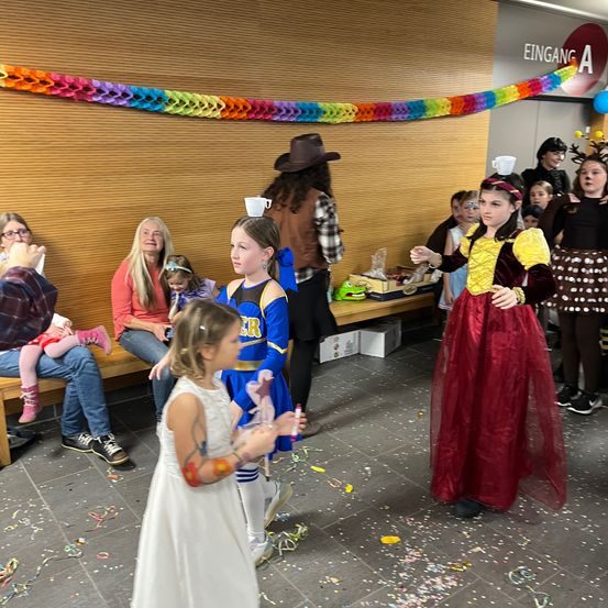 Mehrere Kinder führen auf einer Bühne auf, eines balanciert einen Becher auf dem Kopf. Ein Publikum, bestehend aus Erwachsenen und Kindern, beobachtet sie von den Bänken aus. Bunte Dekorationen schmücken die Wand.