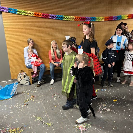 Eine Gruppe von Kindern in verschiedenen Kostümen steht zusammen, während Erwachsene auf einer Bank sitzen. Eine bunte Girlande hängt an der Wand und der Boden ist mit Konfetti übersät.