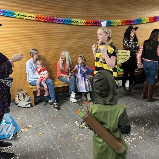 Mehrere Erwachsene und Kinder sitzen auf einer Bank, wobei zwei junge Mädchen stehen. Ein Mädchen ist als Biene gekleidet und das andere trägt ein grünes Kostüm. Sie scheinen sich in einer Partysituation zu befinden, mit Konfetti, das auf dem Boden verstreut ist.
