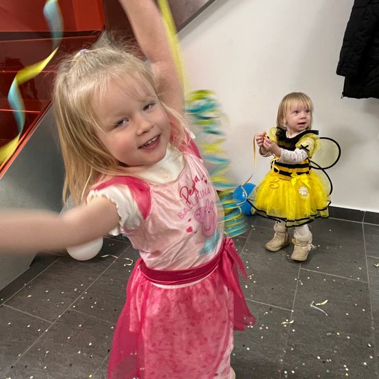 Zwei kleine Mädchen in Kostümen, eines in Pink und eines in Gelb, spielen in einem Raum mit Luftballons und Konfetti auf dem Boden. Dahinter ist eine Treppe und eine weiße Wand zu sehen.