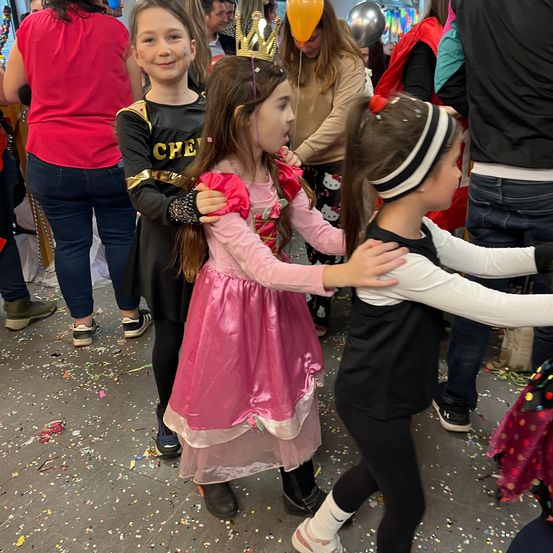 Mehrere Kinder spielen bei einer Party zusammen. Ein Mädchen trägt ein rosa Kleid und eine Krone. Ein anderes Mädchen ist in Schwarz mit einem Stirnband gekleidet. Ein drittes Kind trägt ein Superheldenkostüm. Der Boden ist mit Konfetti bedeckt.