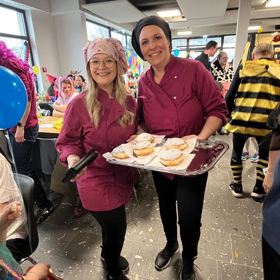 Zwei Frauen in Kochuniformen halten einen Tablett mit Donuts auf einer Party. Sie lächeln und stehen in der Nähe von Tischen, Ballons und Menschen in Kostümen.