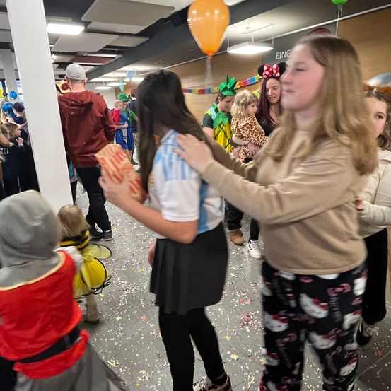 In einem geschäftigen Flur feiern eine Gruppe von Menschen, einige in Kostümen, mit Luftballons und Konfetti. Eine Frau hilft einer anderen mit einem Geschenk, während andere zuschauen.