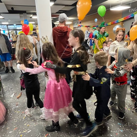 Eine Gruppe von Kindern und Erwachsenen versammelt sich in einem Raum, wahrscheinlich zu einer Feier. Sie tragen Kostüme und einige haben Luftballons. Die Kinder spielen und umarmen sich gegenseitig.
