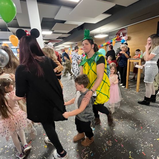Eine Menschenmenge, möglicherweise auf einer Party, mit Luftballons und Konfetti auf dem Boden. Eine Frau in einer Ananas-Kostüm hält ein Kind. Eine andere Frau mit einer Mickey-Mouse-Kopfbedeckung steht in der Nähe.