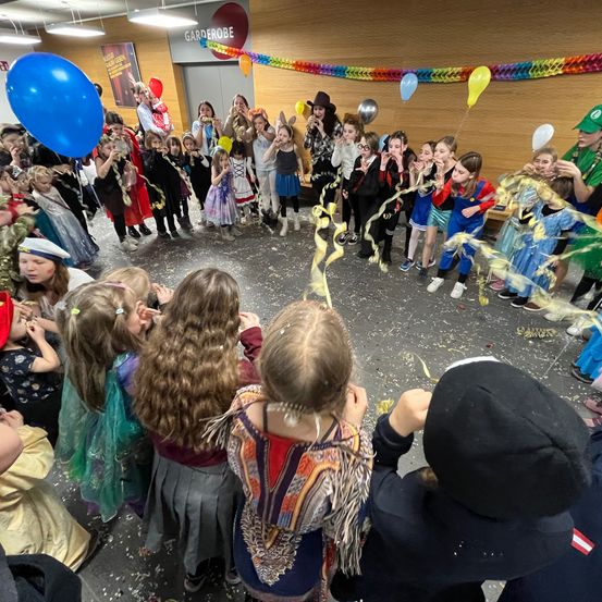 Eine große Gruppe von Kindern in Kostümen, einige blasen Partyhörner, versammelt sich in einem Kreis mit Luftballons und Konfetti in einer festlichen Indoor-Umgebung.