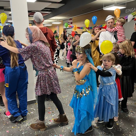 Eine Gruppe von Kindern in Kostümen tanzt in einem Saal mit Konfetti, Luftballons und einem Schild mit der Aufschrift "Eingang". Einige tragen Stiefel, während eines ein weißen Luftballon hält.