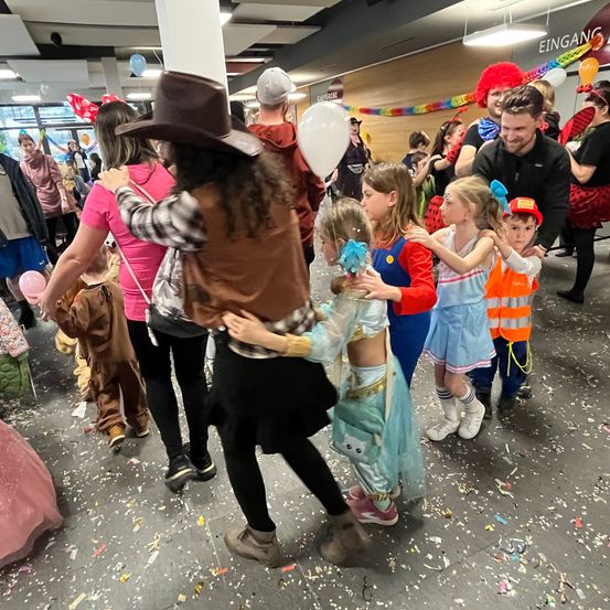 Eine Gruppe von Menschen, darunter Kinder in Kostümen, versammelt sich in einem Flur, möglicherweise für eine Party. Eine Person in einem Cowboyhut hält ein Kind.