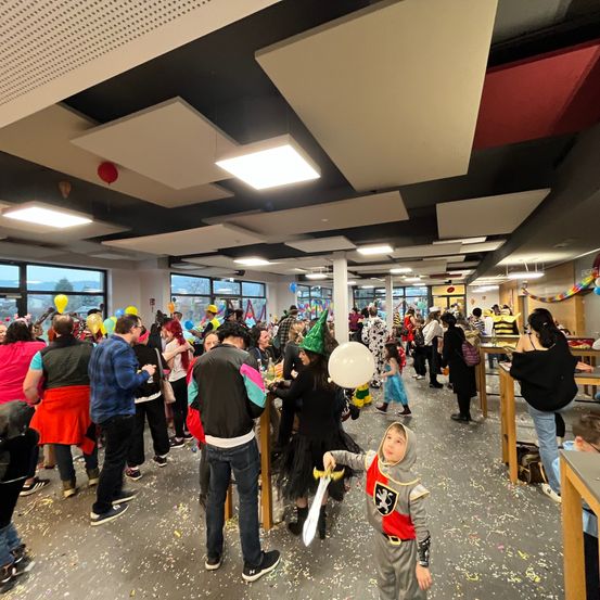 Eine Indoor-Veranstaltung mit vielen Menschen, einige in Kostümen, stehen und sitzen um Tische herum, ein Kind in einem Ritterkostüm mit einem Schwert, Ballons und Luftschlangen hängen von der Decke, Tische mit Gegenständen und ein weißer Ballon auf dem Boden.