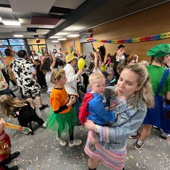 Eine Gruppe von Menschen feiert in einem farbenfrohen Raum. Sie tragen Kostüme und einige halten Luftballons. Eine Frau hält ein Baby in ihren Armen.