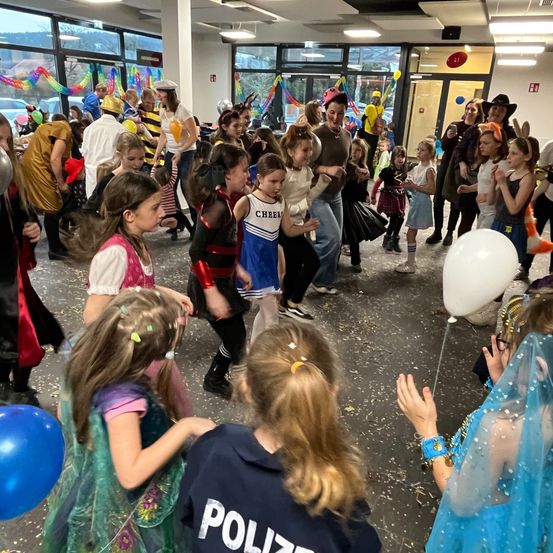 In einem überfüllten Raum tanzen und spielen zahlreiche Kinder in verschiedenen Kostümen. Konfetti fällt um sie herum, und Ballons schweben in der Luft. Ein Polizeibeamter ist ebenfalls unter den Kindern anwesend.