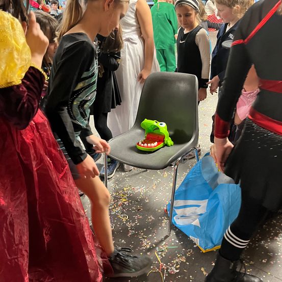 Eine Gruppe von Kindern versammelt sich um einen Stuhl mit einer grünen Spielzeugfigur eines Monsters. Sie sind in verschiedenen Kostümen gekleidet, darunter ein Mädchen in einem roten Kleid und ein Junge in einem Superheldenkostüm.
