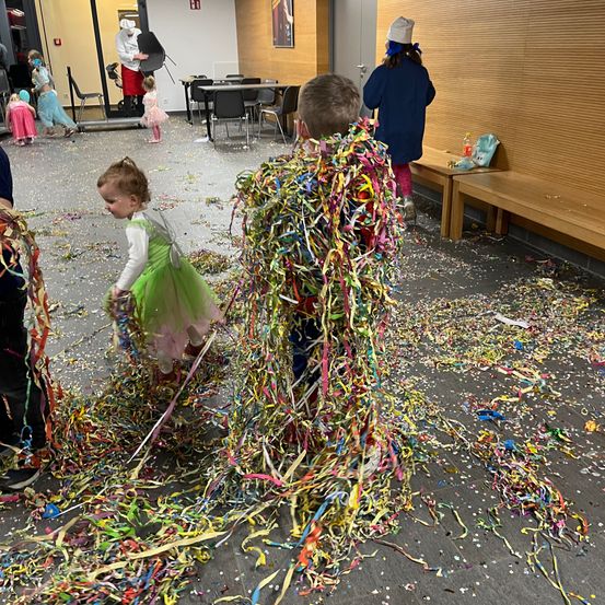 Eine Gruppe von Kindern spielt in einem mit bunten Konfetti bedeckten Raum. Ein Kind hält einen Haufen bunter Bänder.