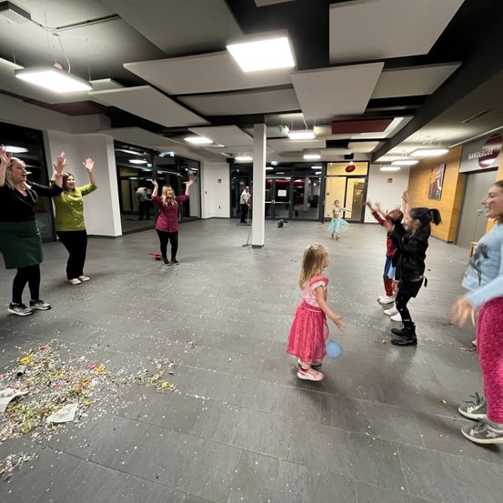 Eine Gruppe von Erwachsenen und Kindern tanzt in einem großen Raum mit Konfetti auf dem Boden. Einige halten Luftballons, und ein Mädchen in einem rosa Kleid trägt einen blauen Ballon.