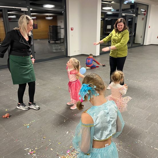 Zwei Frauen und mehrere kleine Kinder spielen in einem geräumigen Raum mit gefliestem Boden. Ein Kind trägt ein Spider-Man-Kostüm und scheint auf dem Boden zu krabbeln. Die Frauen lächeln und scheinen an einer spielerischen Aktivität beteiligt zu sein.