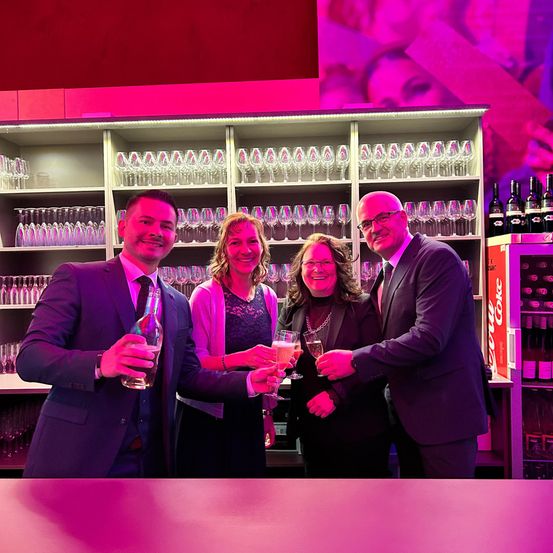 Vier Personen in Anzug und Abendkleid stehen lächelnd hinter einer Bar und machen ein Foto. Sie halten Gläser mit Wein und eine Flasche in den Händen. Hinter ihnen befinden sich Regale mit Gläsern und Flaschen, rechts ist ein Coca-Cola-Schild zu sehen.