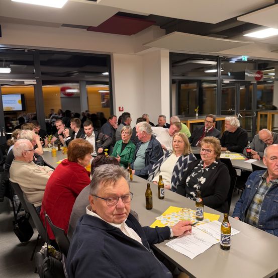 Eine Gruppe älterer Menschen sitzt an einem Tisch, wahrscheinlich in einem Restaurant. Sie haben Flaschen, Papiere und Gläser. Der Raum hat Glaswände und Türen und eine Decke mit Lichtern.